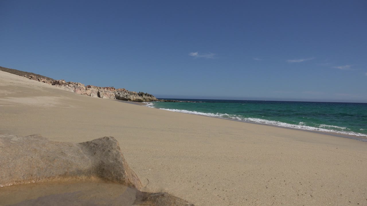A Los Cabos, Mexico shoreline frame shows small white waves along the sand, the foreground rock near the lower edge, pale rocky dunes to the left, and no visible people.