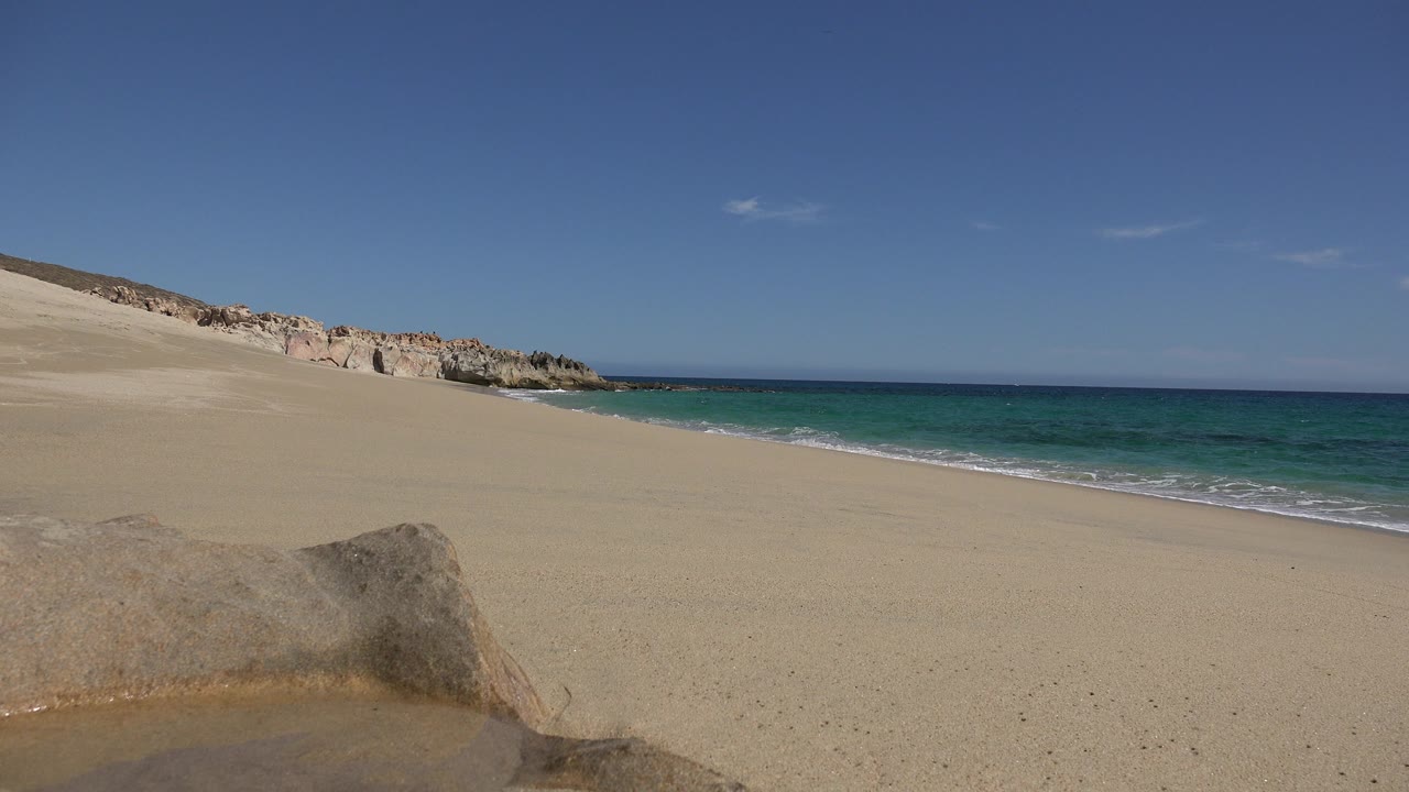 Los Cabos beach view at the start of the clip, with pale sand, a foreground rock, turquoise water, a rocky wall in the distance, and no visible people.