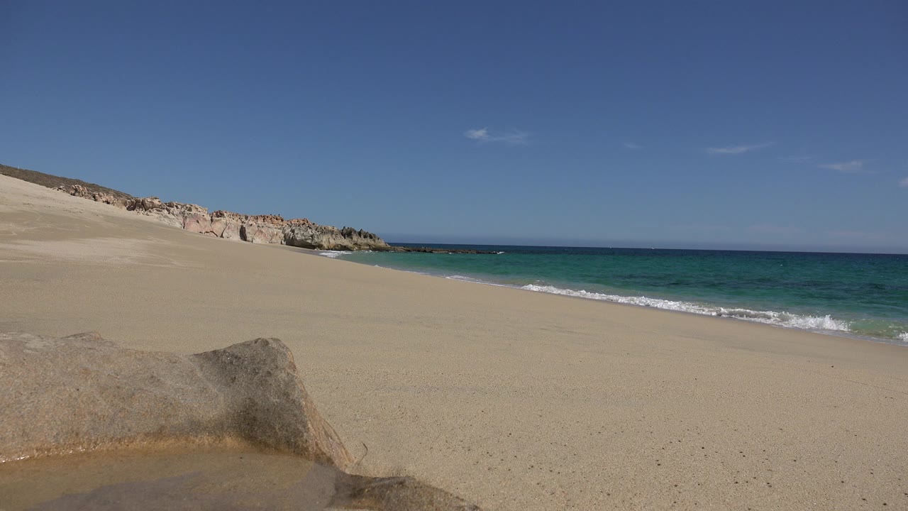 Wide view of a Los Cabos sandy beach with a foreground rock, low dunes to the left, blue ocean to the right, and no visible people.