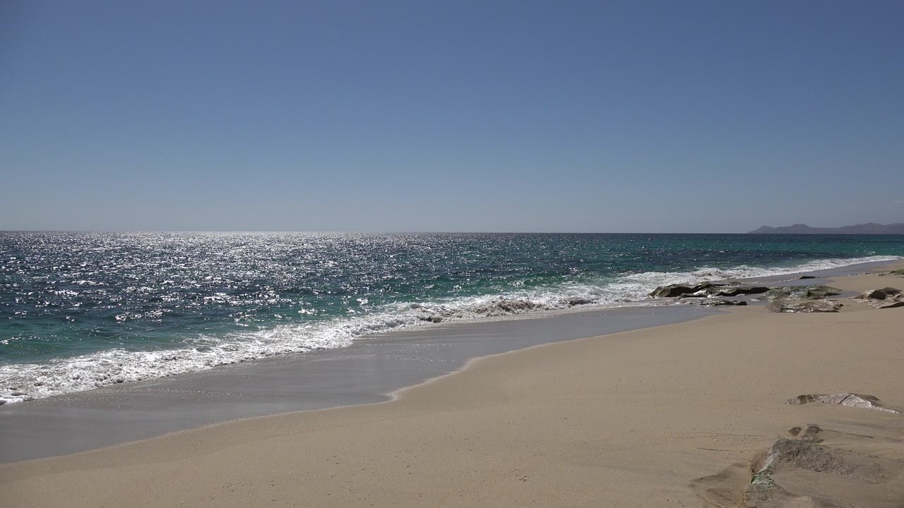 Opening view of Los Cabos surf washing onto pale sand, with rocks near the right edge, distant land on the horizon, and no visible people.