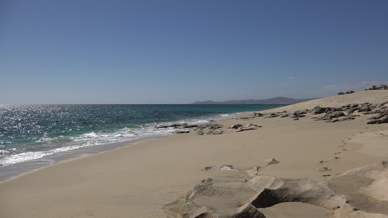 Opening frame of a Los Cabos, Mexico beach with small waves touching the left shoreline, tan sand and rocks in the foreground, distant mountains, and no visible people.