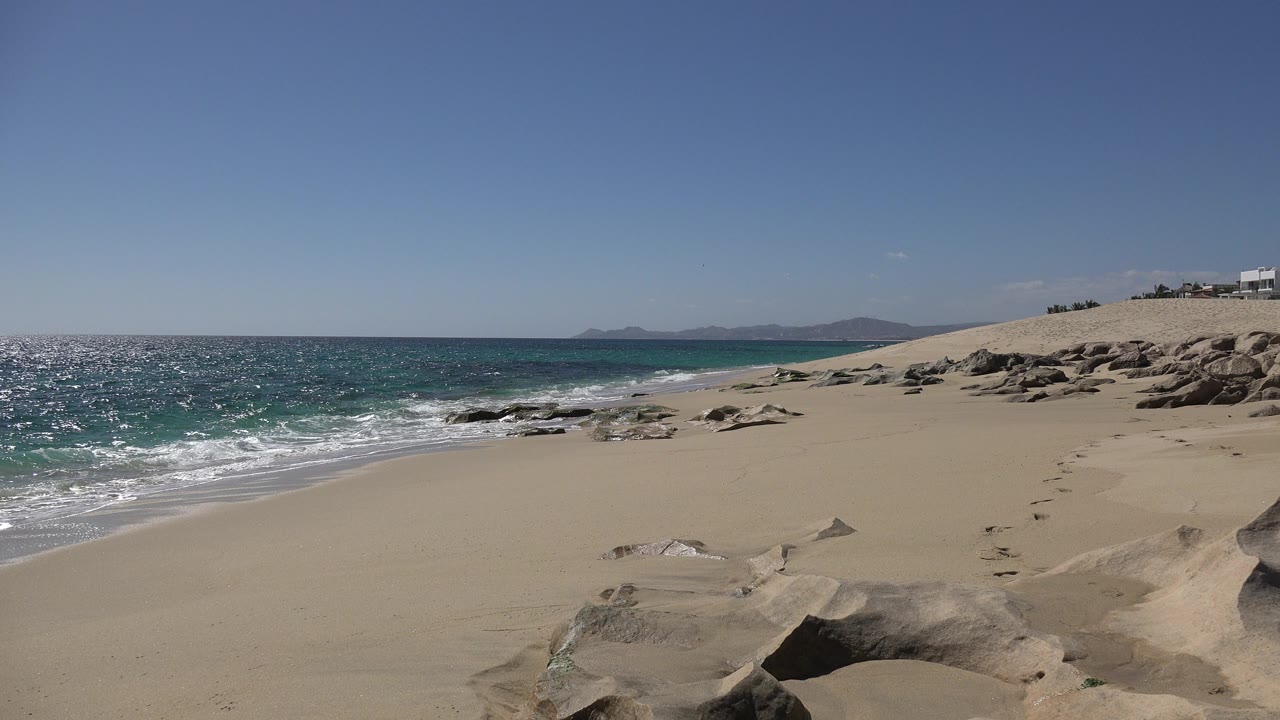 Empty beach with breaking waves, rock formations, and distant hills.