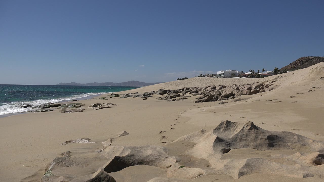 Los Cabos beach view with dark foreground rocks, pale sand, turquoise surf on the left, dunes on the right, distant mountains, and no visible people.