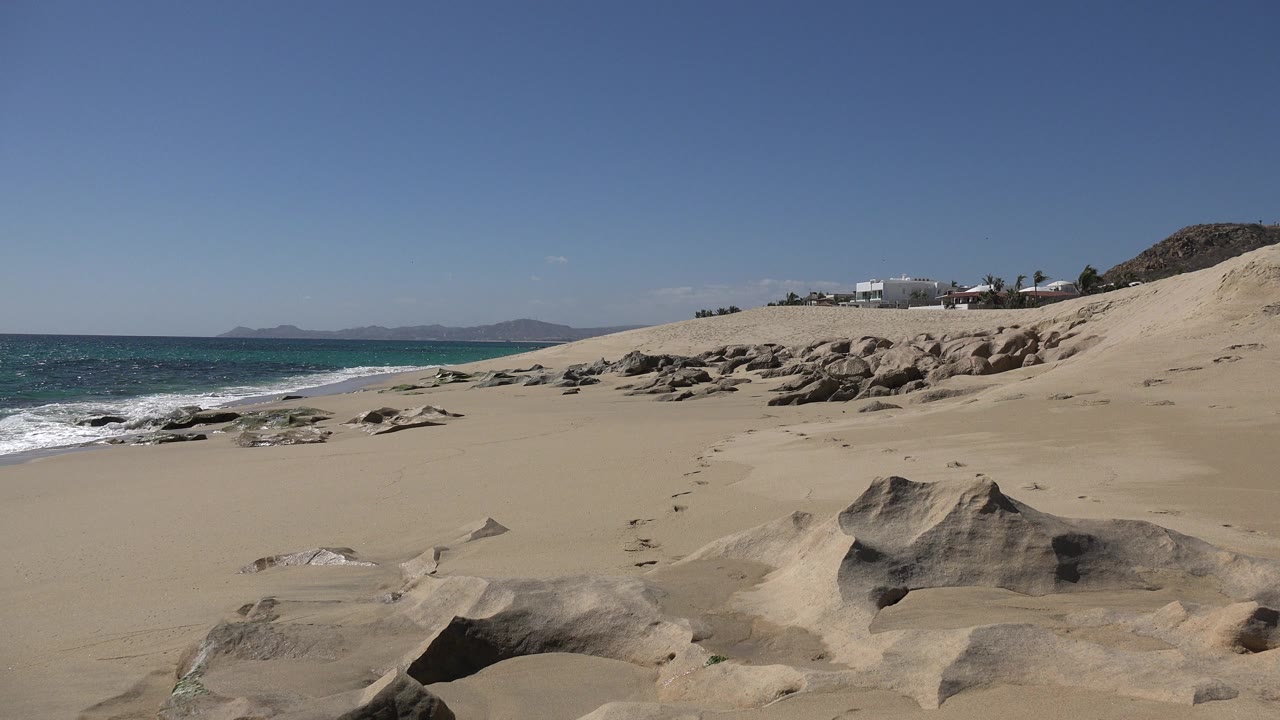Los Cabos shoreline frame with broad empty sand, rounded foreground rock, breaking surf at left, dune slope at right, distant buildings, and no visible people.