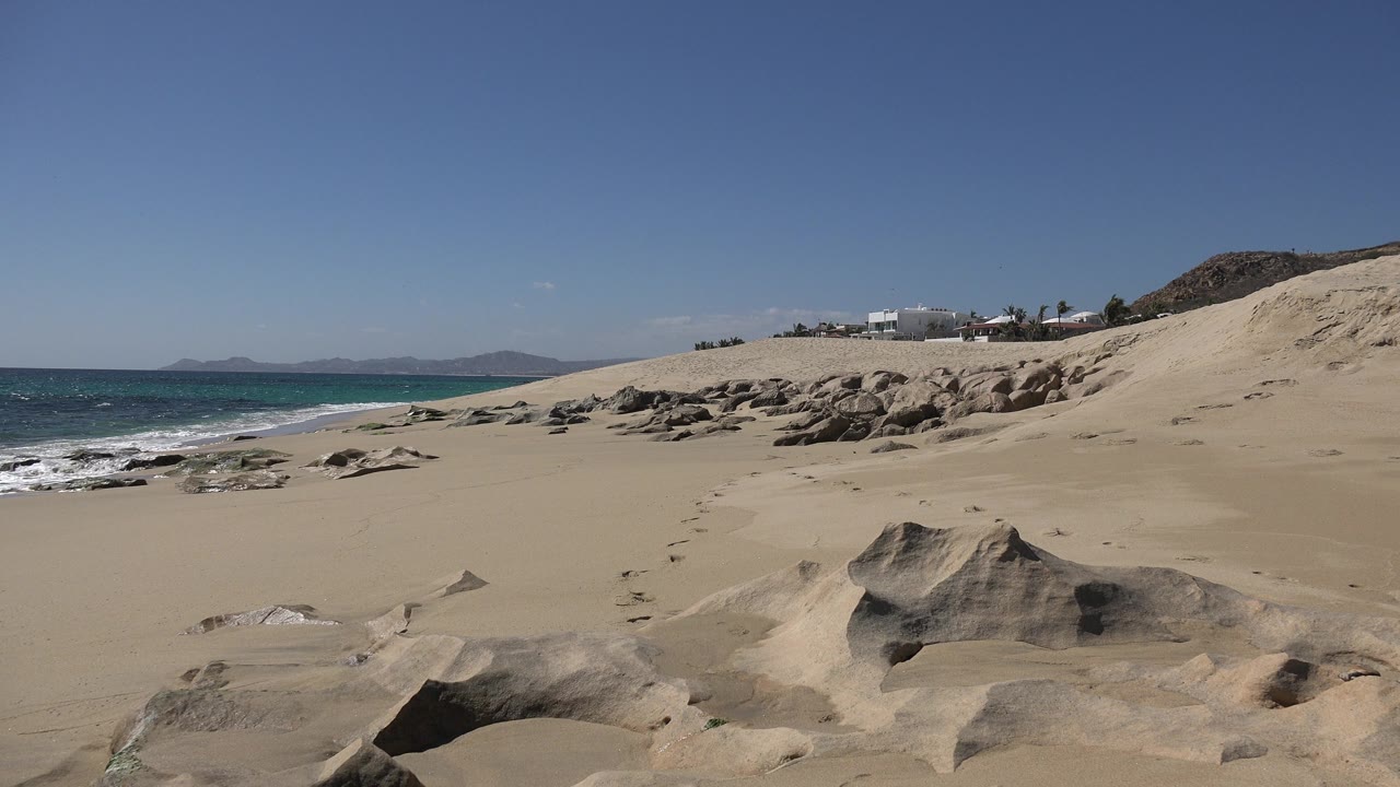 Wide sandy shoreline with boulders, ocean, dunes, and blue sky.
