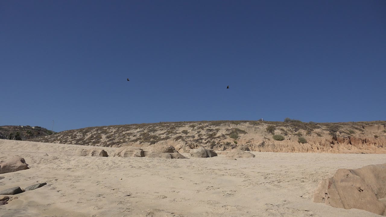 Los Cabos, Mexico frame showing pale sandy terrain, scattered rounded rocks, a low scrub-covered ridge, deep blue sky, and no visible people.