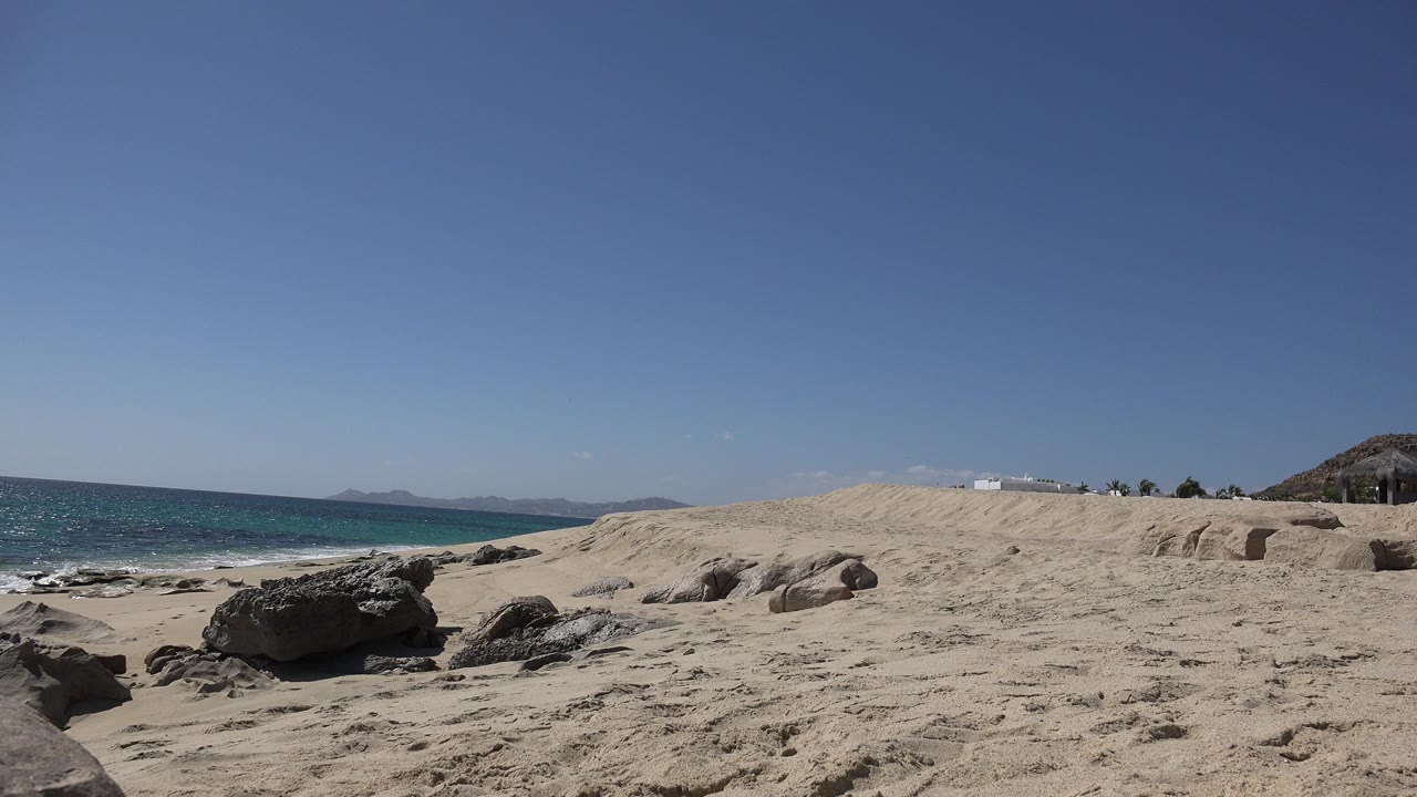 Opening frame of a Los Cabos sandy beach with dark shoreline rocks, turquoise ocean to the left, a dry rocky rise to the right, and no visible people.