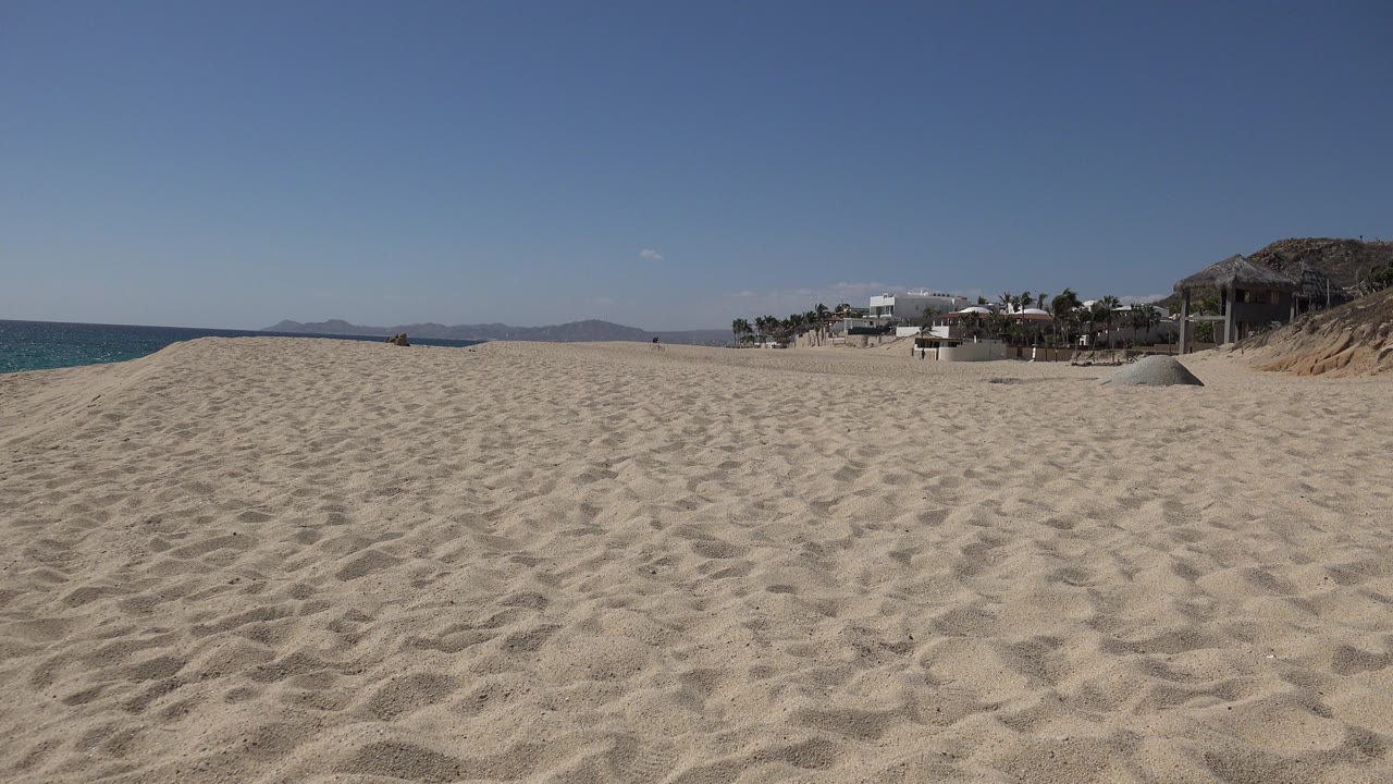 Opening view of a Los Cabos beach with rippled sand in the foreground, blue ocean along the left edge, distant hills, shoreline houses to the right, and no visible people.