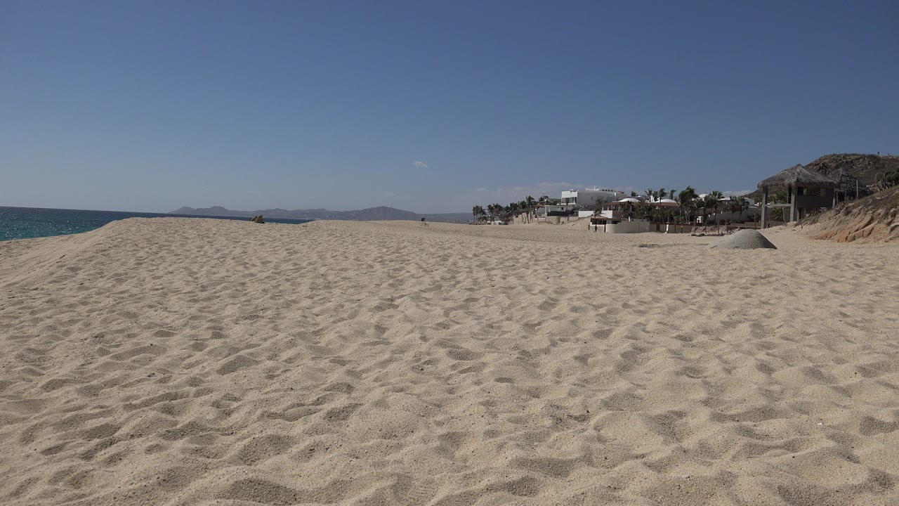 One second into the Los Cabos clip, the sandy foreground remains dominant while the ocean sits left, hazy hills cross the horizon, and distant beach houses remain on the right with no visible people.