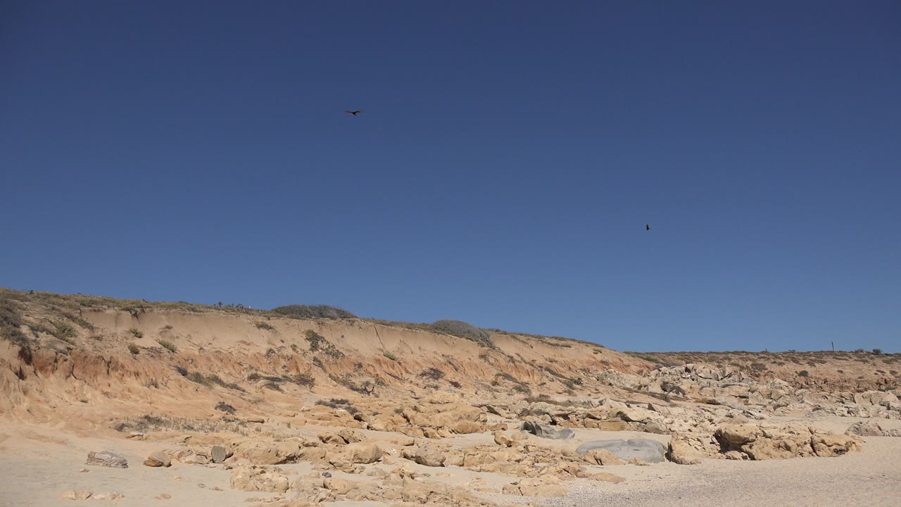 A wide cloudless sky dominates the frame while tan arid hillside, rocks, and scrub sit low in Los Cabos, Mexico, with no visible people.