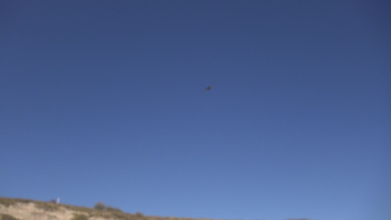At 0:00 in Los Cabos, Mexico, a small dark flying object appears high in clear blue sky above a low tan ridge, with no visible people.