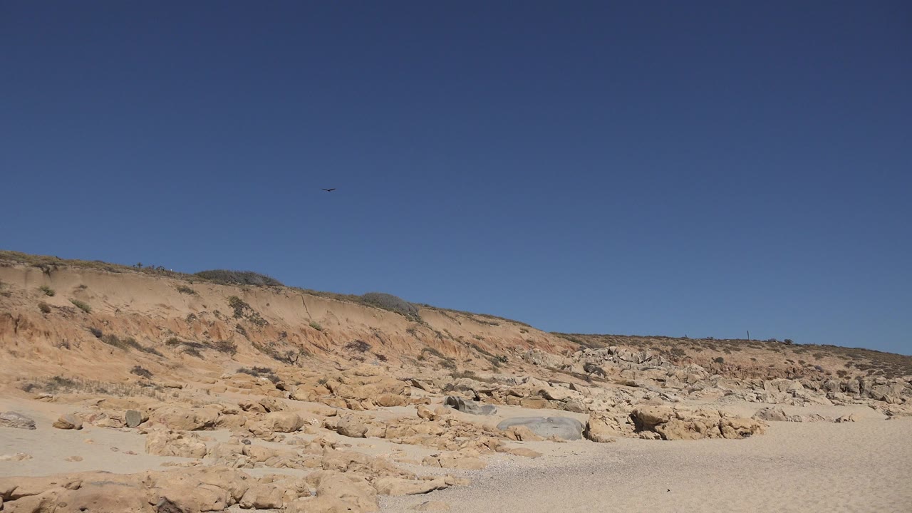 Two small birds are visible high in a clear blue sky above sandy rocks and eroded slopes in Los Cabos, Mexico, with no visible people.