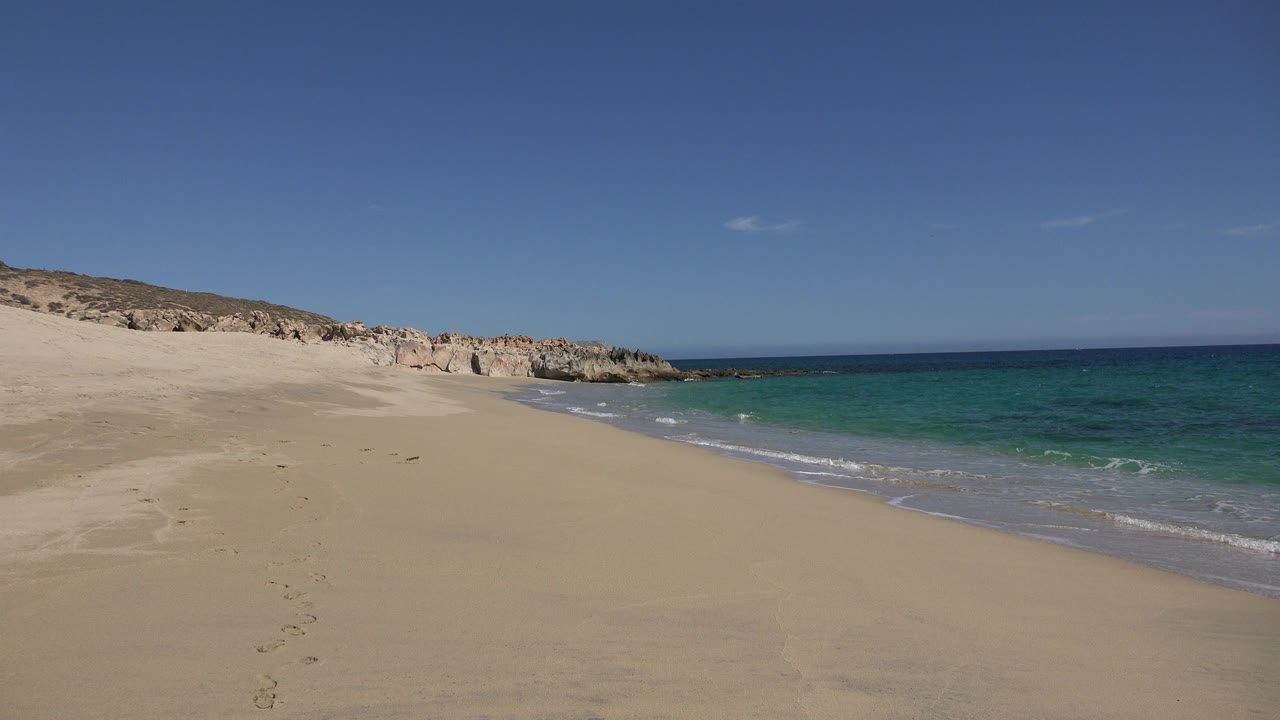 Opening frame of a Los Cabos beach with pale sand in the foreground, blue sea to the right, rocky bluffs in the distance, and no visible people.