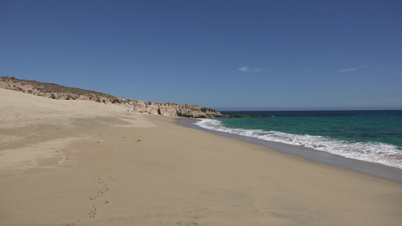 Wide view of a Los Cabos beach with pale sand in front, blue-green water at right, rocky dune edge at left, clear sky above, and no visible people.