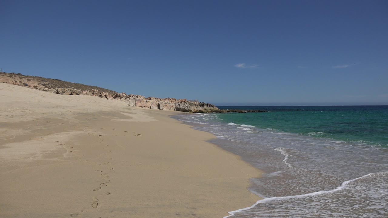 Los Cabos beach at 0:00 with dry sand in the foreground, small surf washing in from the right, rocky dunes on the left, and no visible people.