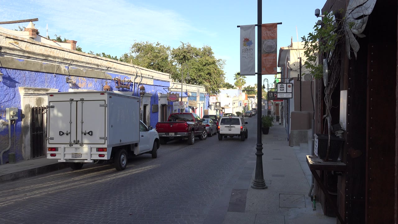 Los Cabos neighborhood street one second in, with the white truck still prominent, a red pickup beside it, shaded right foreground, and no clearly visible people.