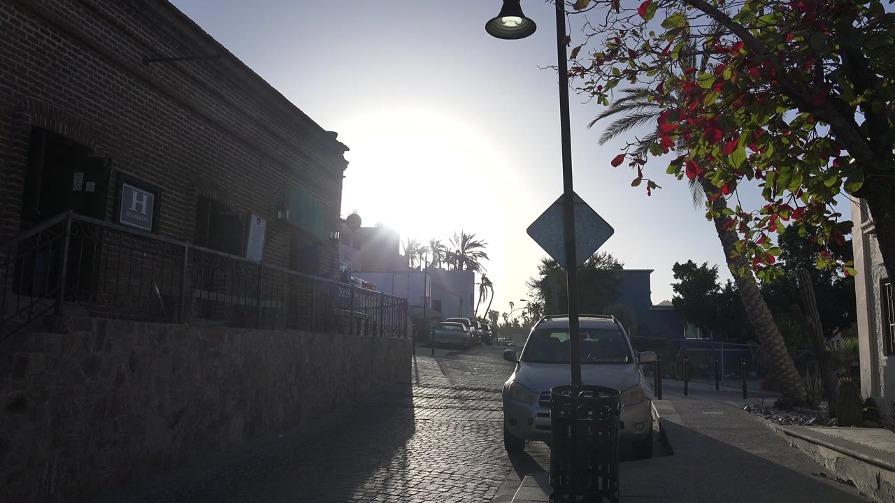 Opening frame in Los Cabos, Mexico, showing a parked car near a brick building, a flowering tree, a street sign, bright sun flare, and no visible people.