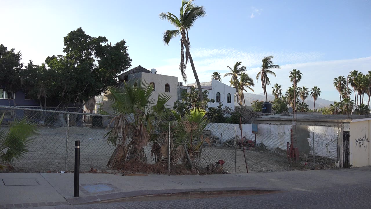 Wide street-level view in Los Cabos, Mexico, with a curb, black bollard, chain-link fence, palms, low residential buildings, distant hills, blue sky, and no visible people.