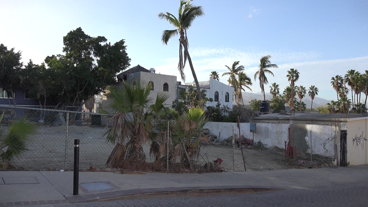 Los Cabos neighborhood view with a foreground curb and bollard, fenced sandy lot, white stucco building, tall palms, hazy hills, clear sky, and no visible people.
