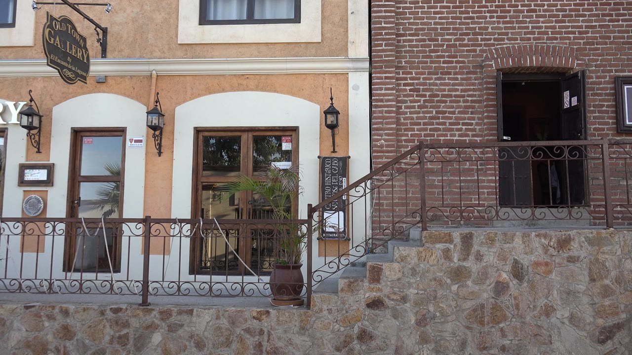 A Los Cabos storefront facade shows the hanging gallery sign above glass doors, a brick side wall, wrought iron railings, stone steps, and no visible people.