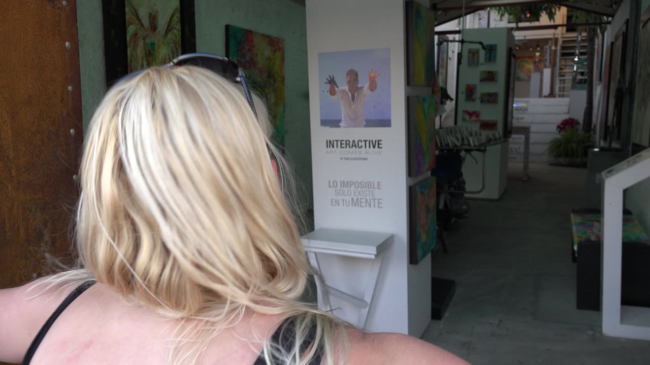 Blonde woman seen from behind in Los Cabos, Mexico, with sunglasses on her head, wall art, a white interactive sign, and a shaded walkway ahead.