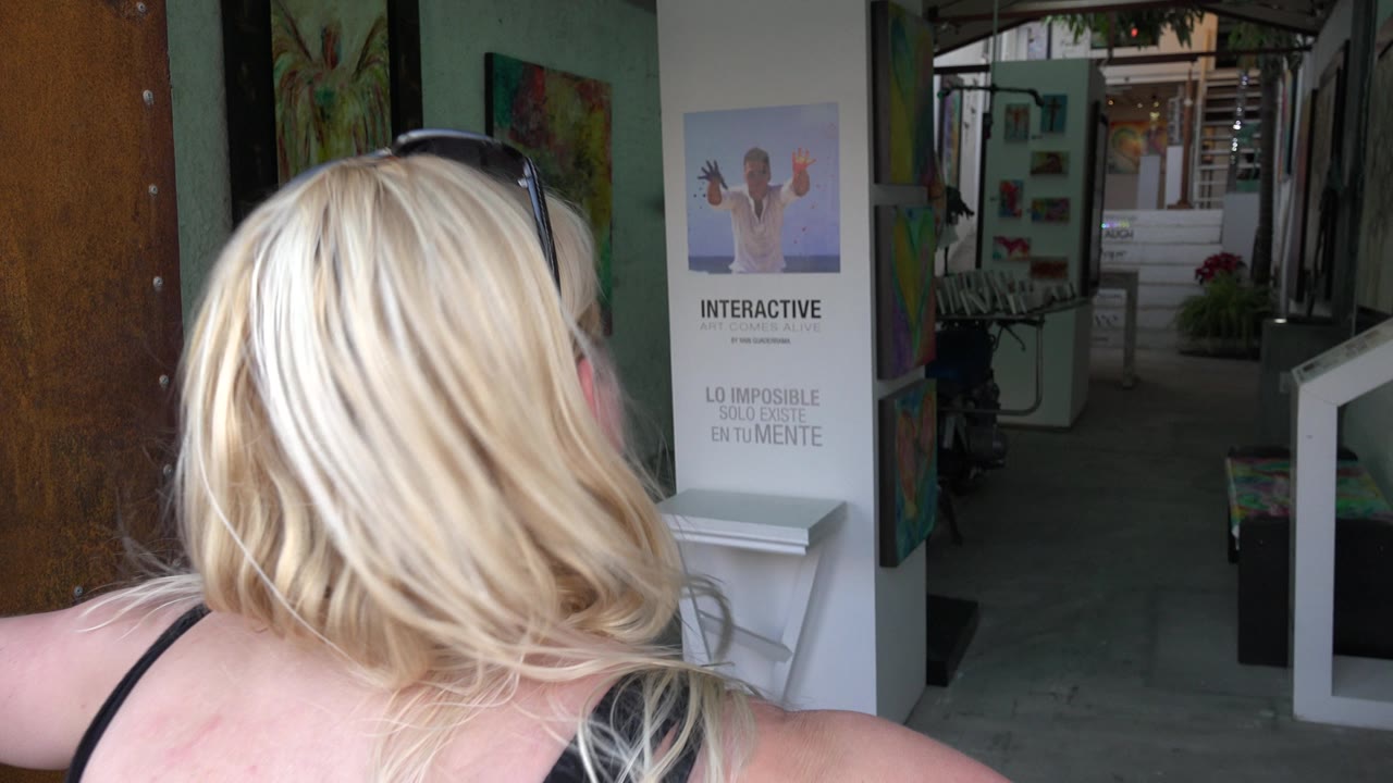 In Los Cabos, Mexico, the woman's hair and shoulder occupy the foreground while paintings, a freestanding interactive sign, and a dim corridor remain visible ahead.