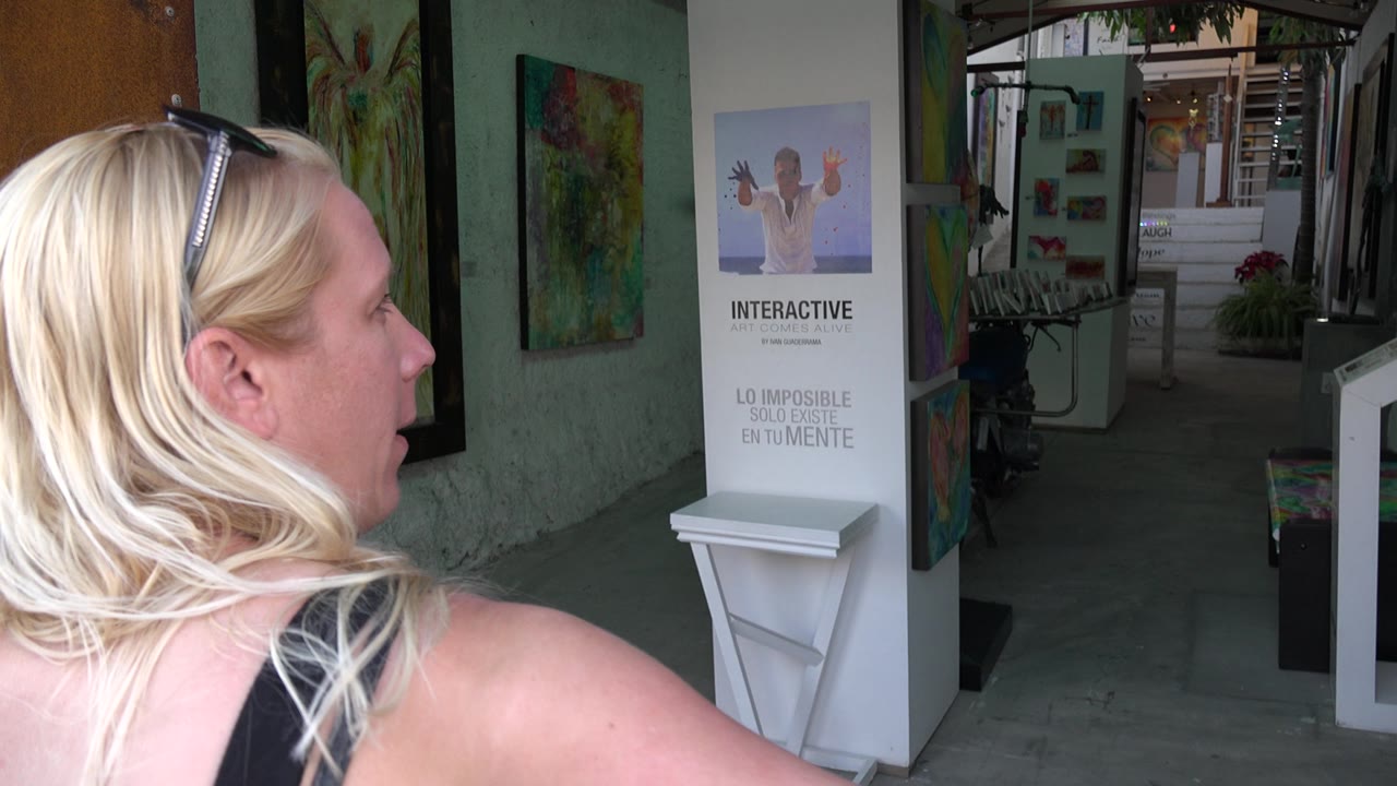 Woman turns in profile near a doorway with gallery paintings and stairs behind her.