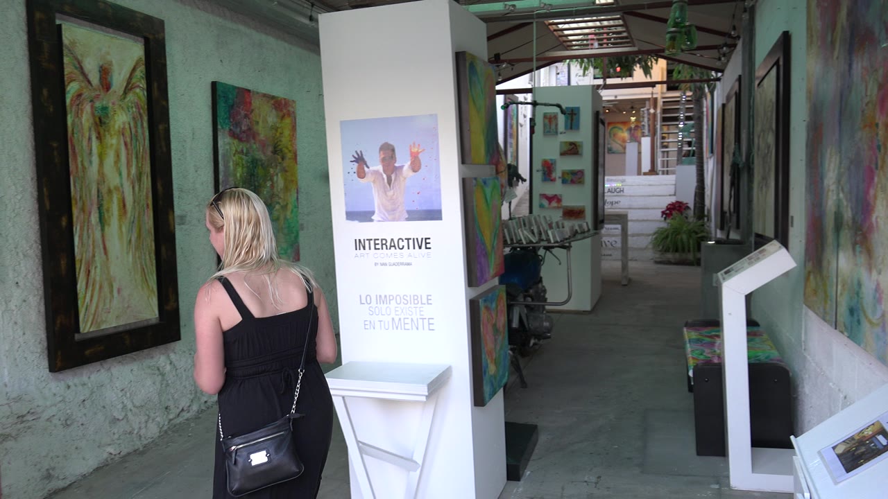 In Los Cabos, Mexico, a blonde woman in a black dress walks beside a mint-colored gallery wall with framed paintings and a white interactive display ahead.
