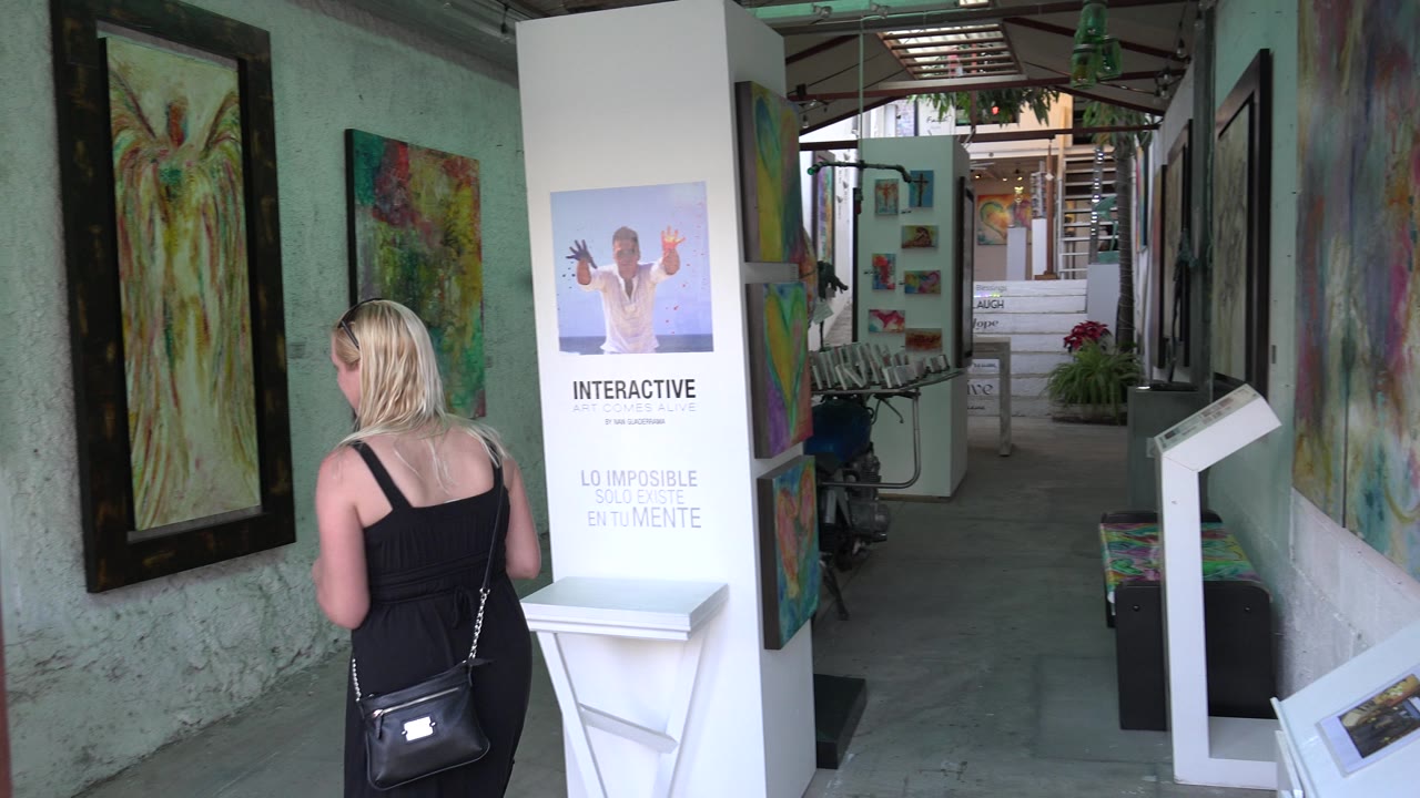The visitor stands closer to the framed painting while a tall white interactive display and more artwork line the narrow Los Cabos gallery walkway.