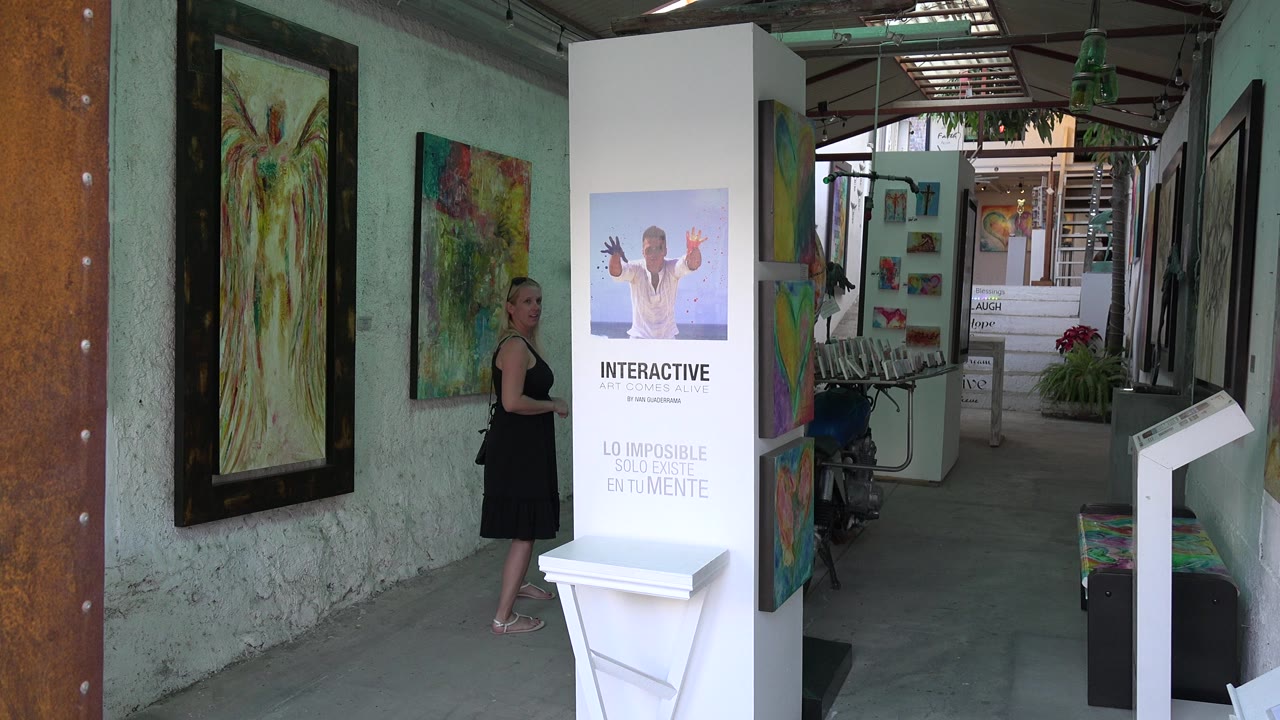 Woman in a black dress standing beside abstract paintings in a gallery corridor.