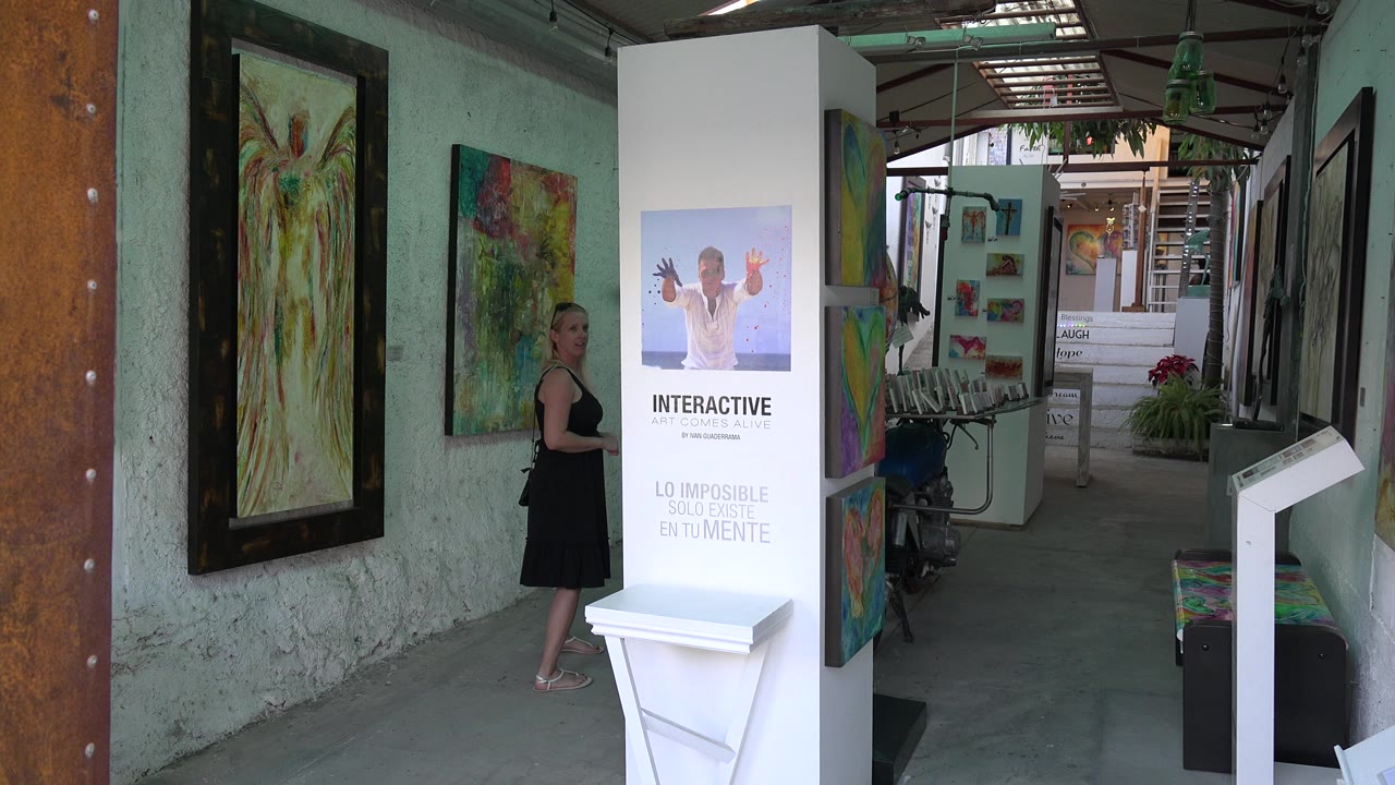 The Los Cabos gallery view holds on the woman near colorful wall art, with the white interactive display stand more centered in the corridor.