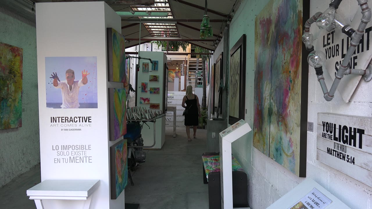 In the Los Cabos hallway, the person remains in the midground while the foreground poster, white pedestal, and colorful paintings frame the narrow walkway.