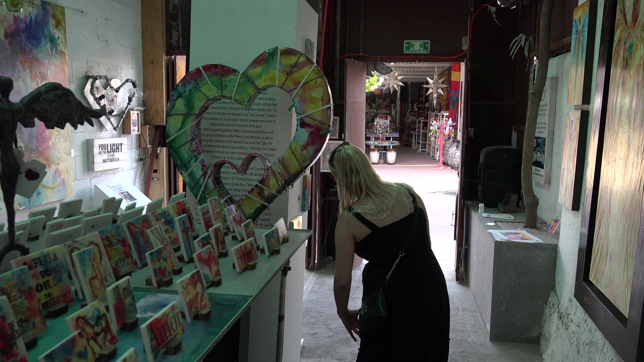 A woman in a black dress leans near a display of colorful cards and paintings inside a Los Cabos indoor art space, with bright doorway light ahead.