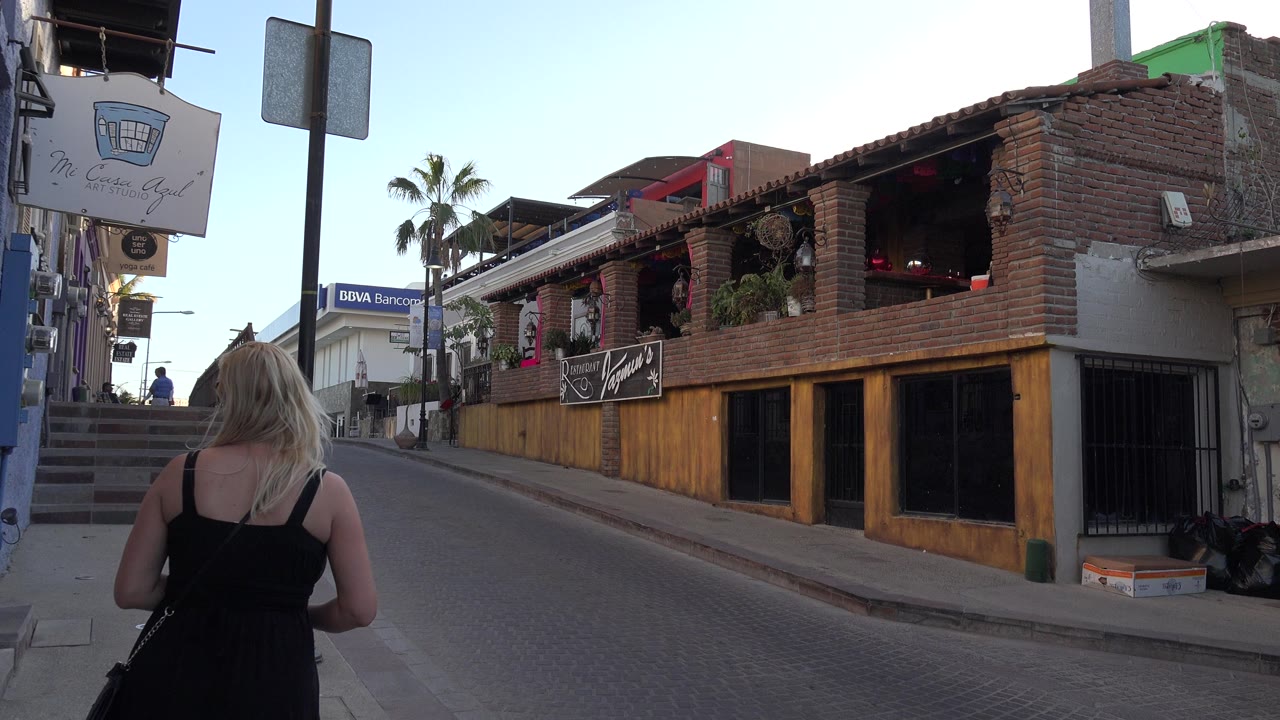 The woman continues away from the camera on the Los Cabos street, with a two-story brick building, hanging plants, and a staircase visible ahead.