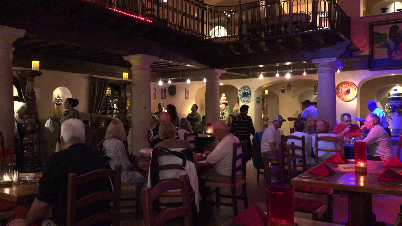 Crowded restaurant interior with diners, red table settings, pillars, and balcony railings.