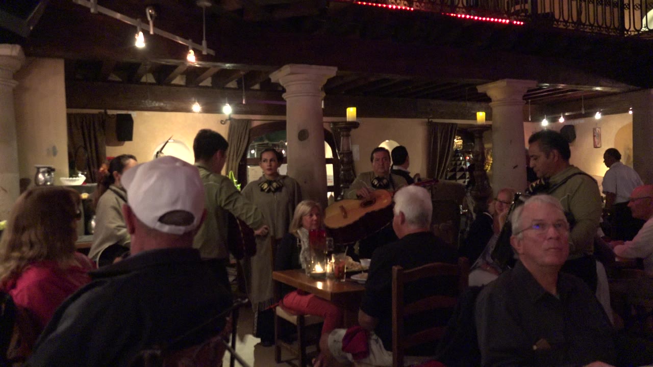 In Los Cabos, musicians in matching outfits stand among seated guests, with a white cap in the foreground and warm overhead lights above the tables.