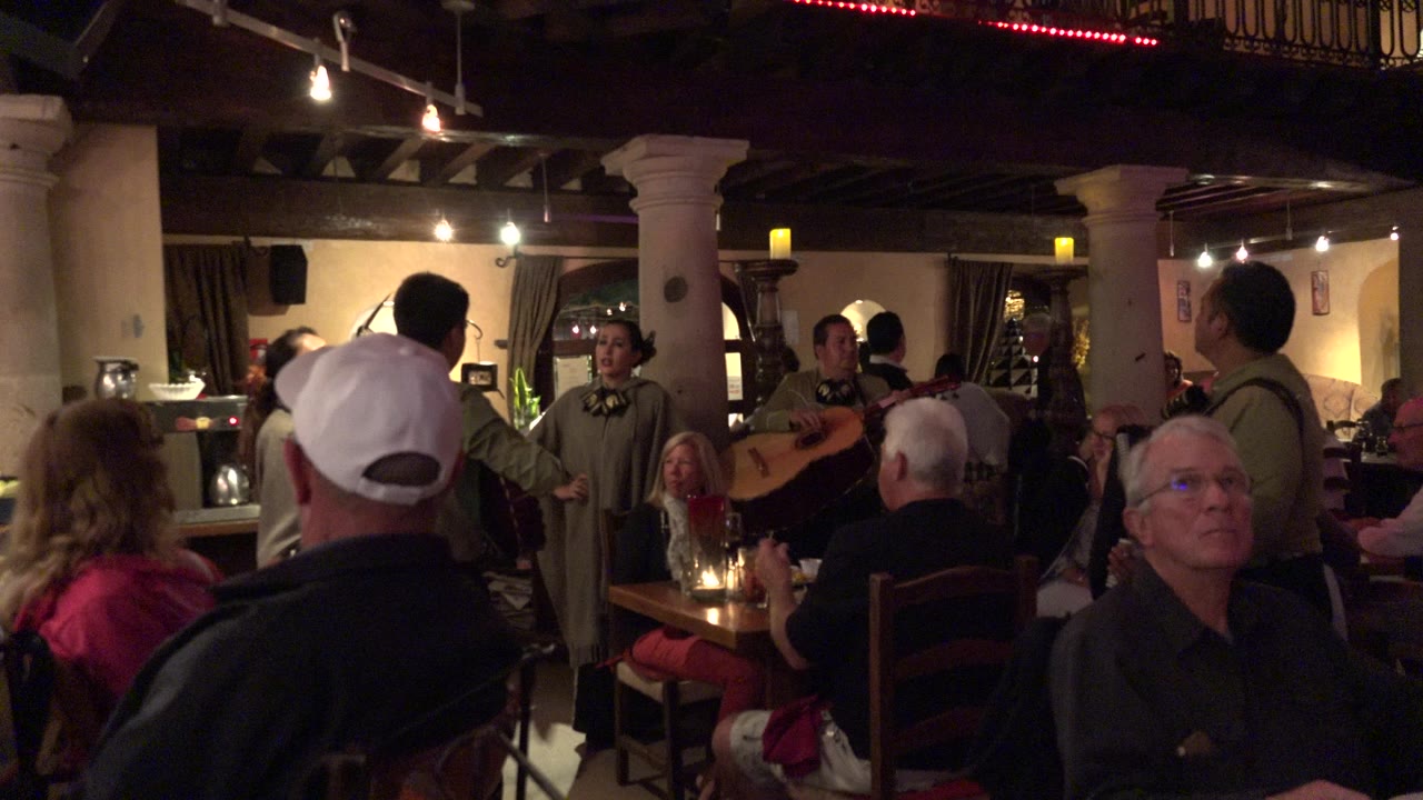 Crowded Los Cabos restaurant interior at the start of the clip, with seated diners in the foreground and musicians performing between tables under warm ceiling lights.