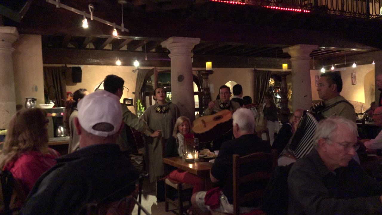 In Los Cabos, Mexico, diners sit close together in a warmly lit restaurant while a singer and musicians perform near the tables at the start of the clip.