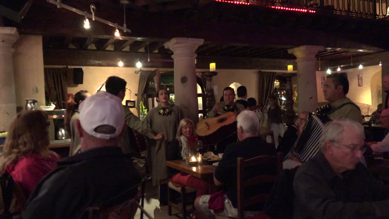 A woman stands near the center of the Los Cabos restaurant while guitar and accordion players face diners seated around small tables with drinks.