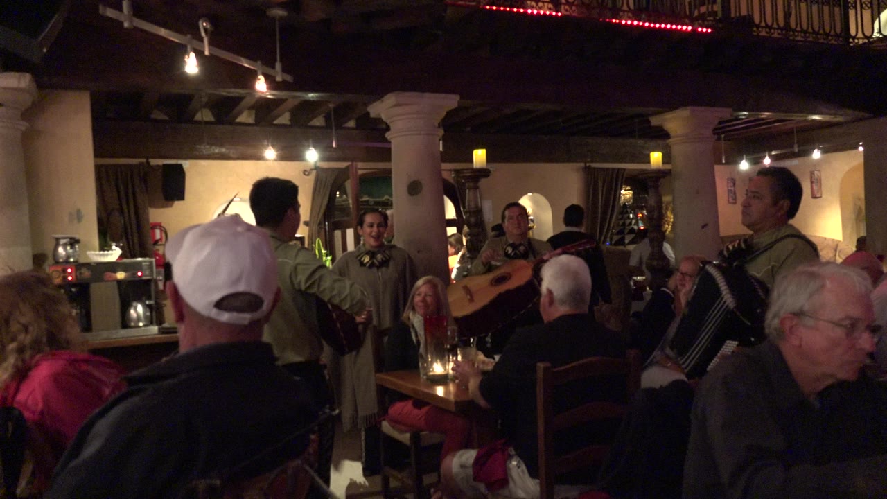 In Los Cabos, Mexico, seated restaurant guests face three costumed musicians performing near tables with candles and glasses under warm ceiling lights.