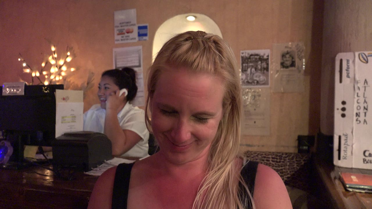 At 0:01 in Los Cabos, Mexico, the foreground blonde woman remains looking down while the background person continues holding a phone beside counter equipment and wall signs.
