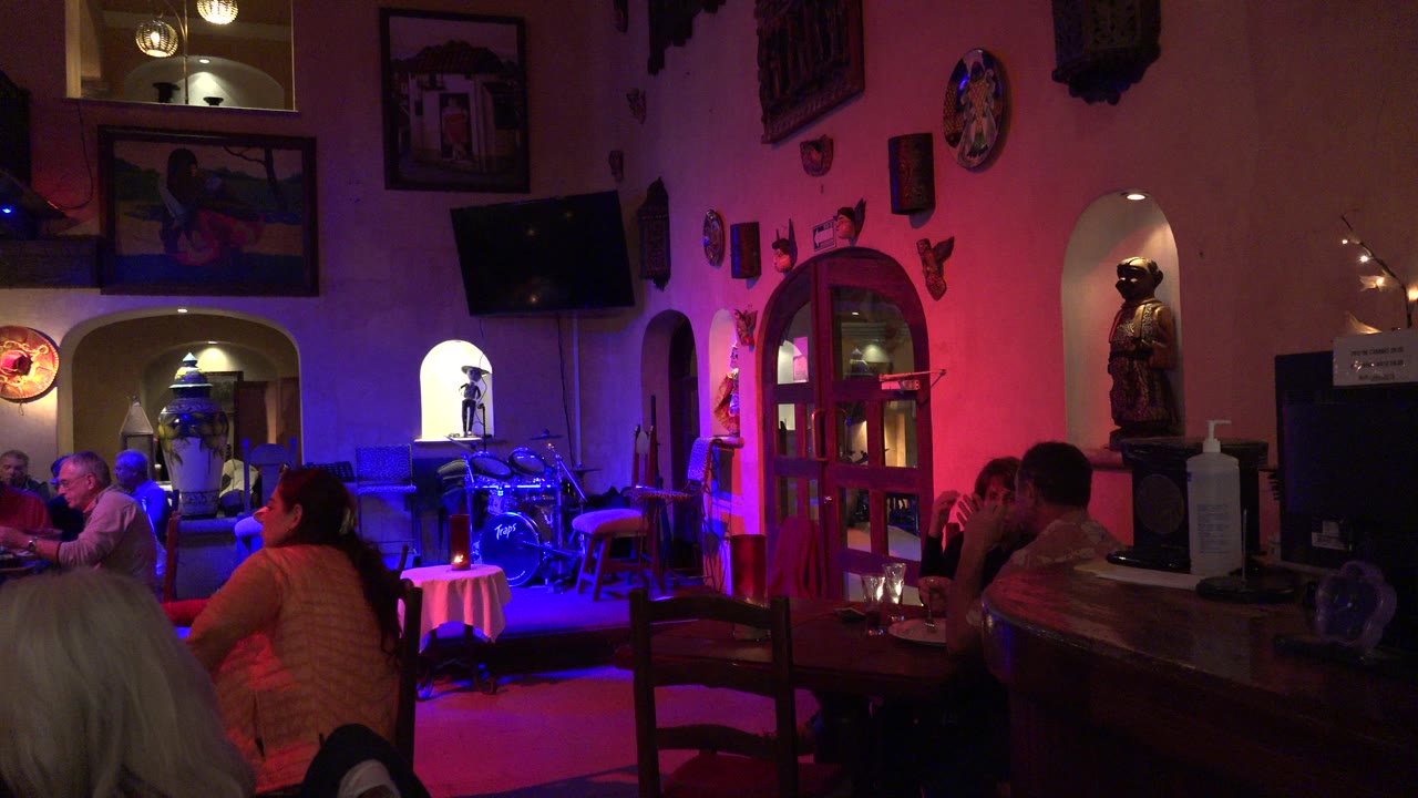Adults sit at tables inside a Los Cabos restaurant as blue floor light and magenta wall light reveal arched niches, framed art, and a red door area.