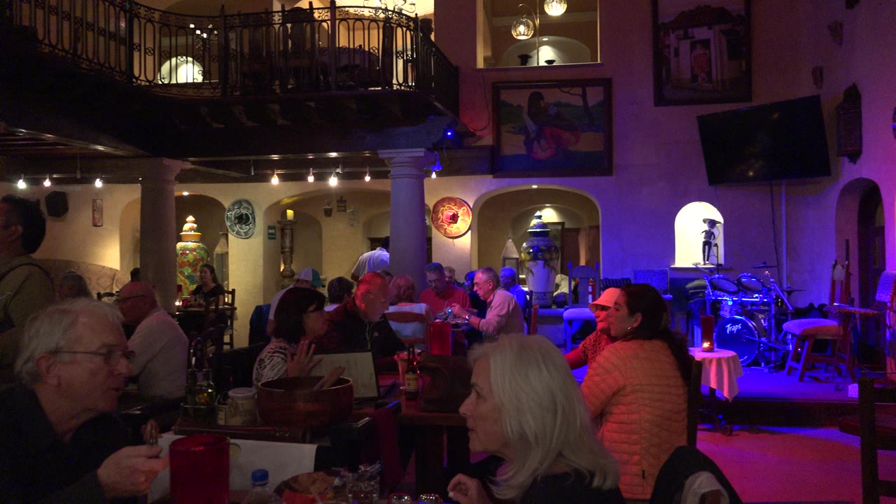 Los Cabos restaurant interior with visible diners at foreground and center tables, purple-blue light on the right, and a balcony above the dining room.