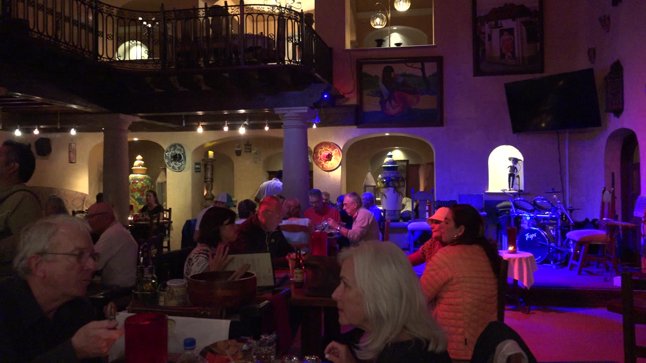 Los Cabos dining room remains crowded with visible people at tables, framed wall art, arched niches, and blue-purple illumination across the right side.
