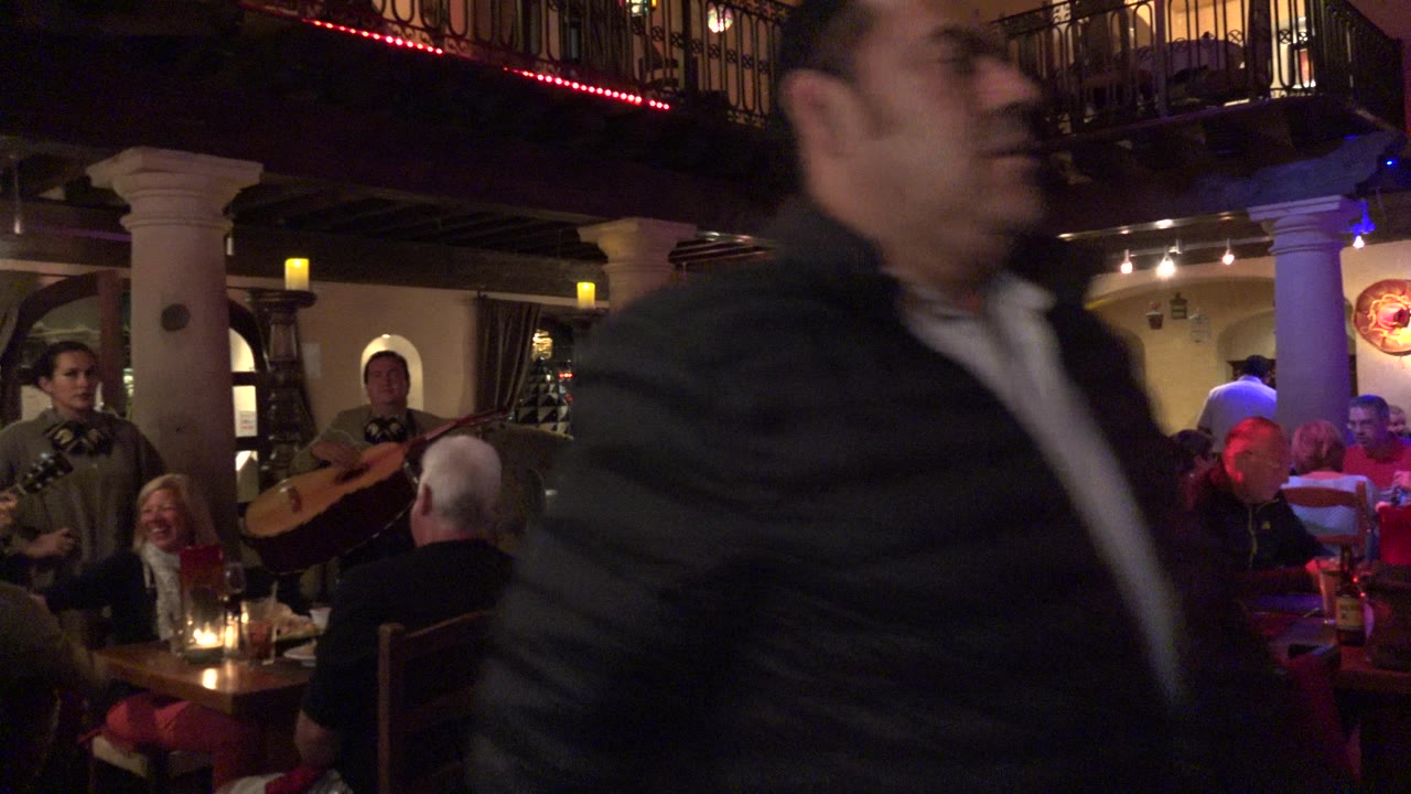 A man moves across the foreground as a mariachi group performs near tables of diners inside the warm-lit Los Cabos restaurant.
