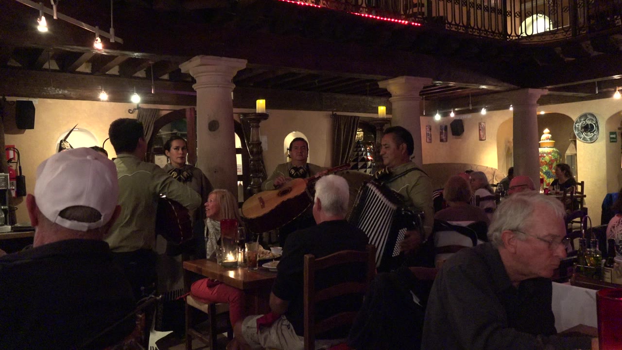 In Los Cabos, Mexico, musicians stand near crowded dining tables while seated guests watch under warm restaurant lighting.