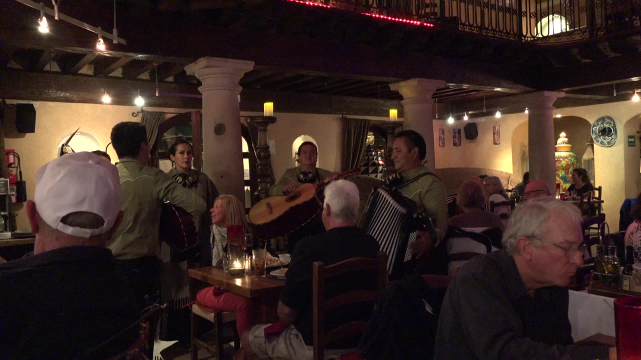 In Los Cabos, Mexico, the performers remain framed by foreground diners, with an accordion player on the right and warm lights across the restaurant interior.