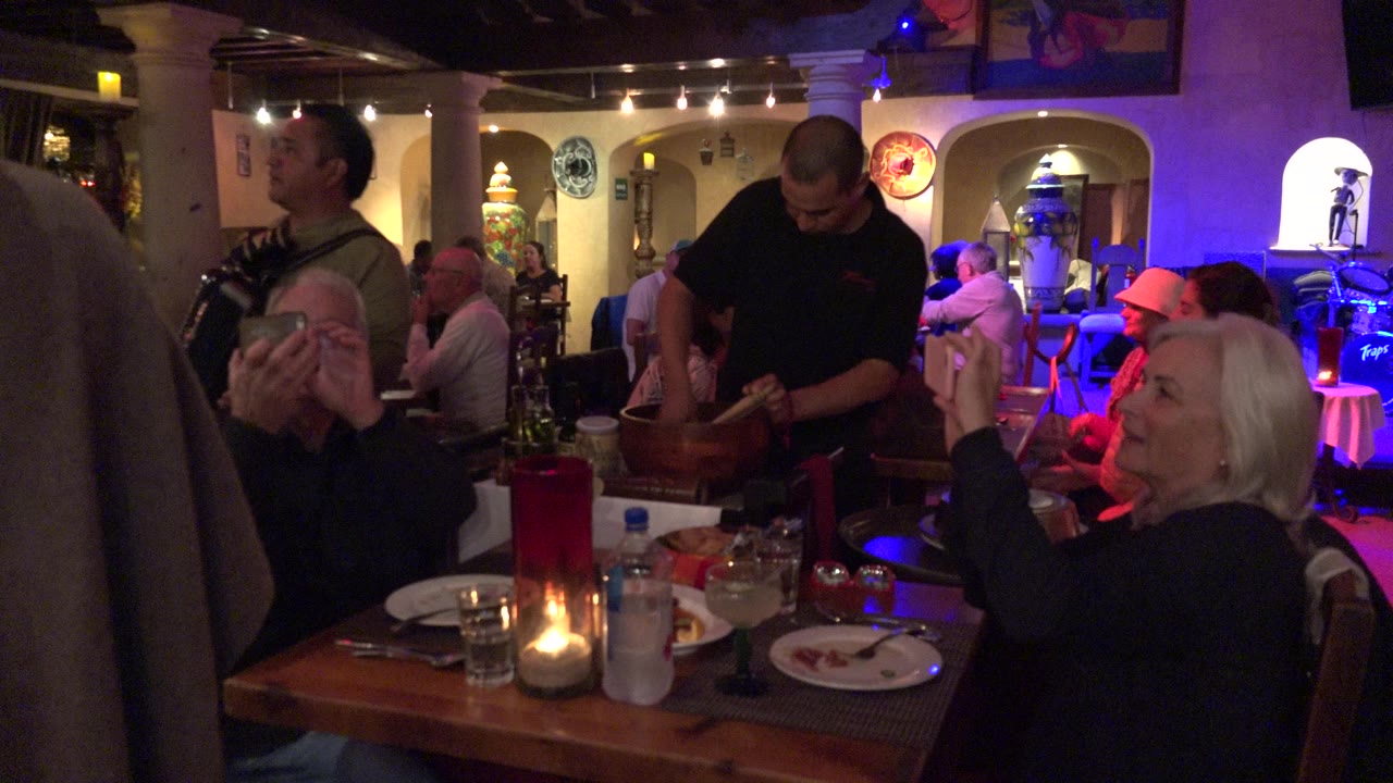 Older diners at a restaurant table while a waiter prepares food nearby.