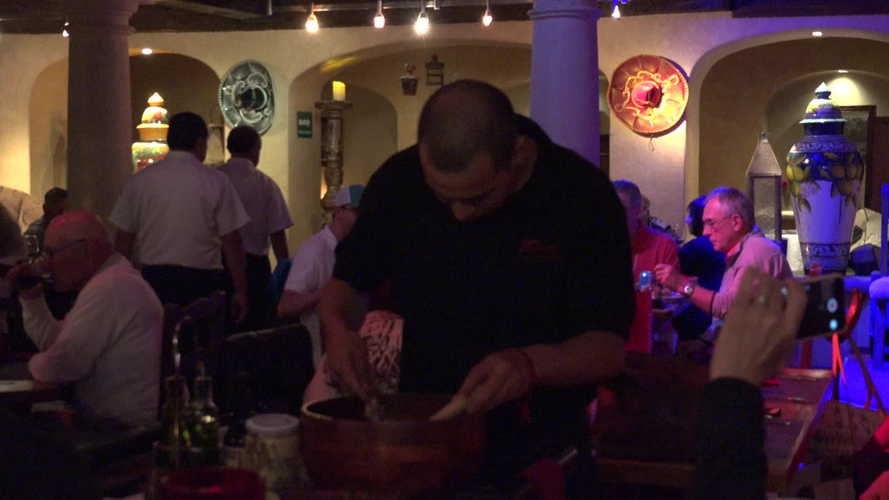 In a Los Cabos restaurant interior, a man in a dark shirt leans over a large bowl while seated diners, arched walls, hanging bulbs, and ceramic decor fill the background.
