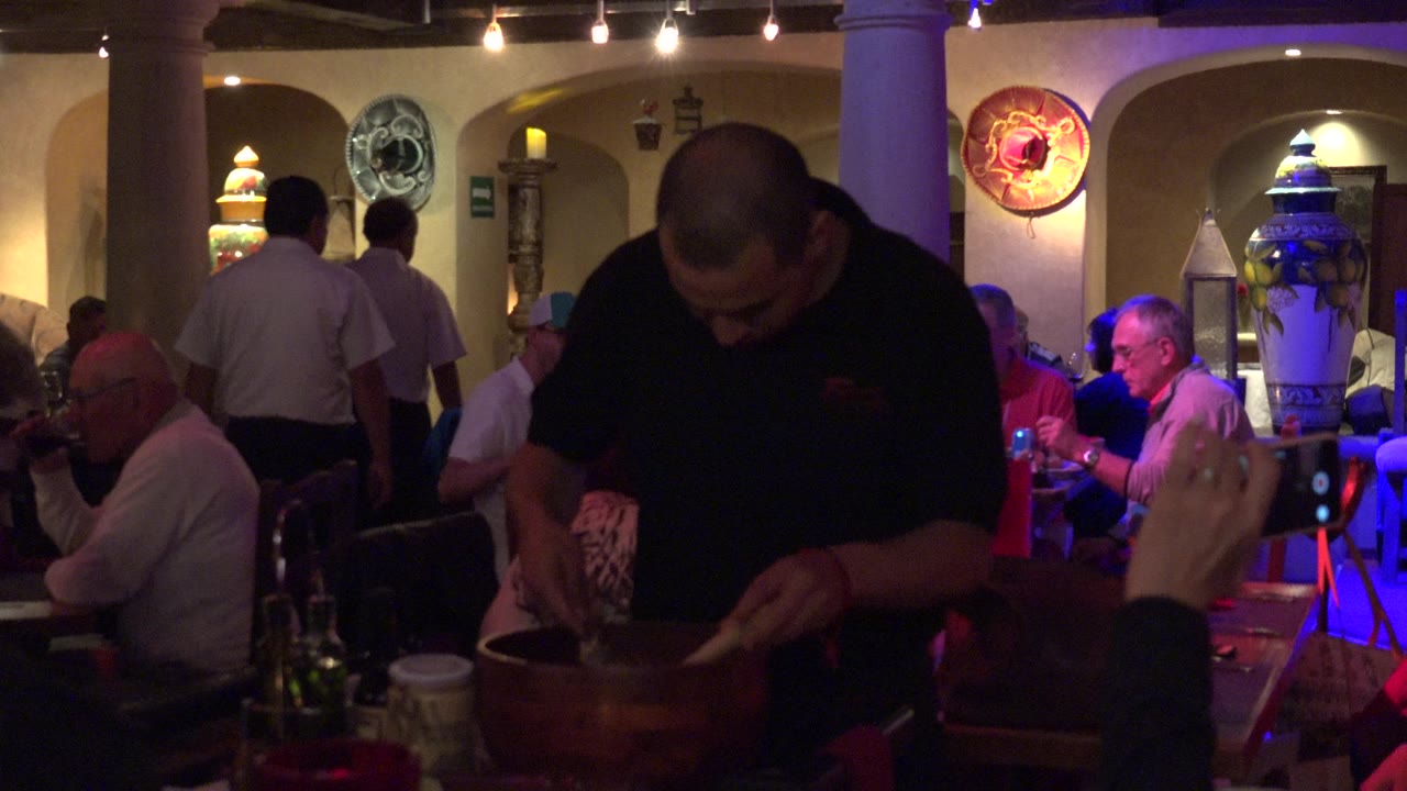 The Los Cabos dining room remains crowded as the foreground worker looks down into the bowl, with a hand holding a phone at the right edge and diners seated under warm and purple lighting.